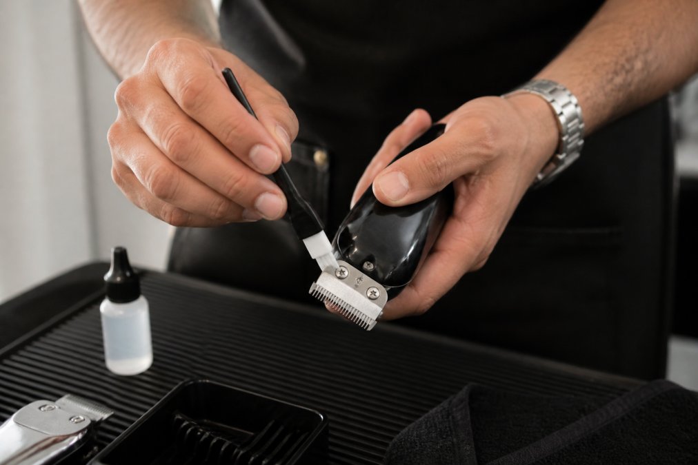 Professional barber's hands applying clipper oil to blade edges with a small brush during maintenance, showing proper technique in natural lighting
