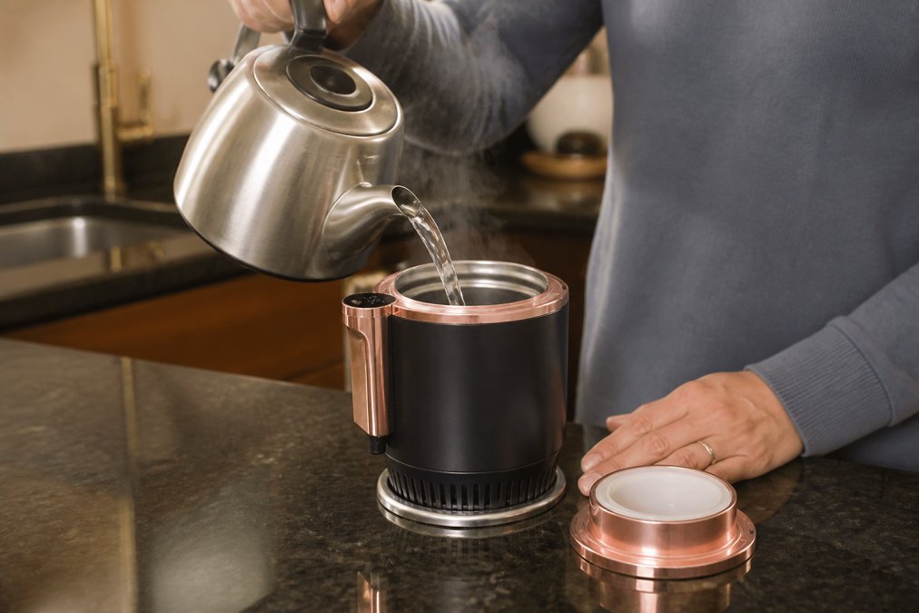 Close-up of hands pouring hot water from a kettle into a stainless steel travel thermos on a kitchen counter, steam rising from the mug, warm kitchen lighting