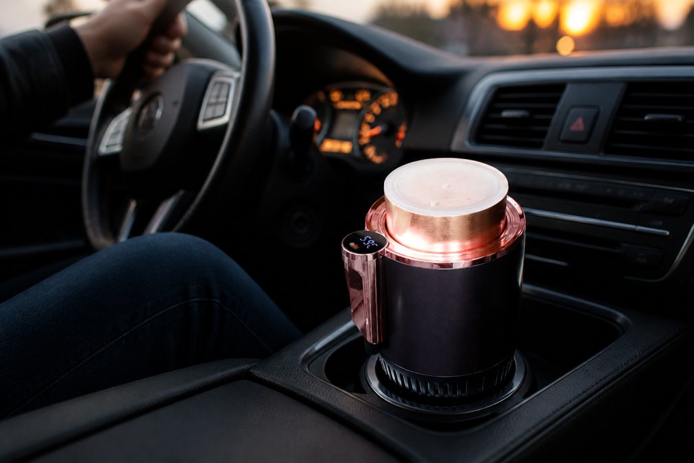 A travel mug sitting in a car cup holder with a digital temperature display glowing blue, dashboard and steering wheel visible in the background during an early morning commute, warm ambient lighting from the instrument cluster