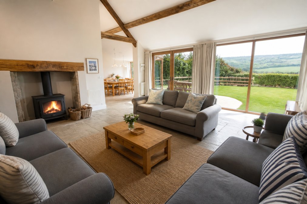 A spacious, bright living area in a holiday barn with comfortable seating, a fireplace, and large windows overlooking countryside, with a fully enclosed garden visible outside, soft natural light streaming through windows