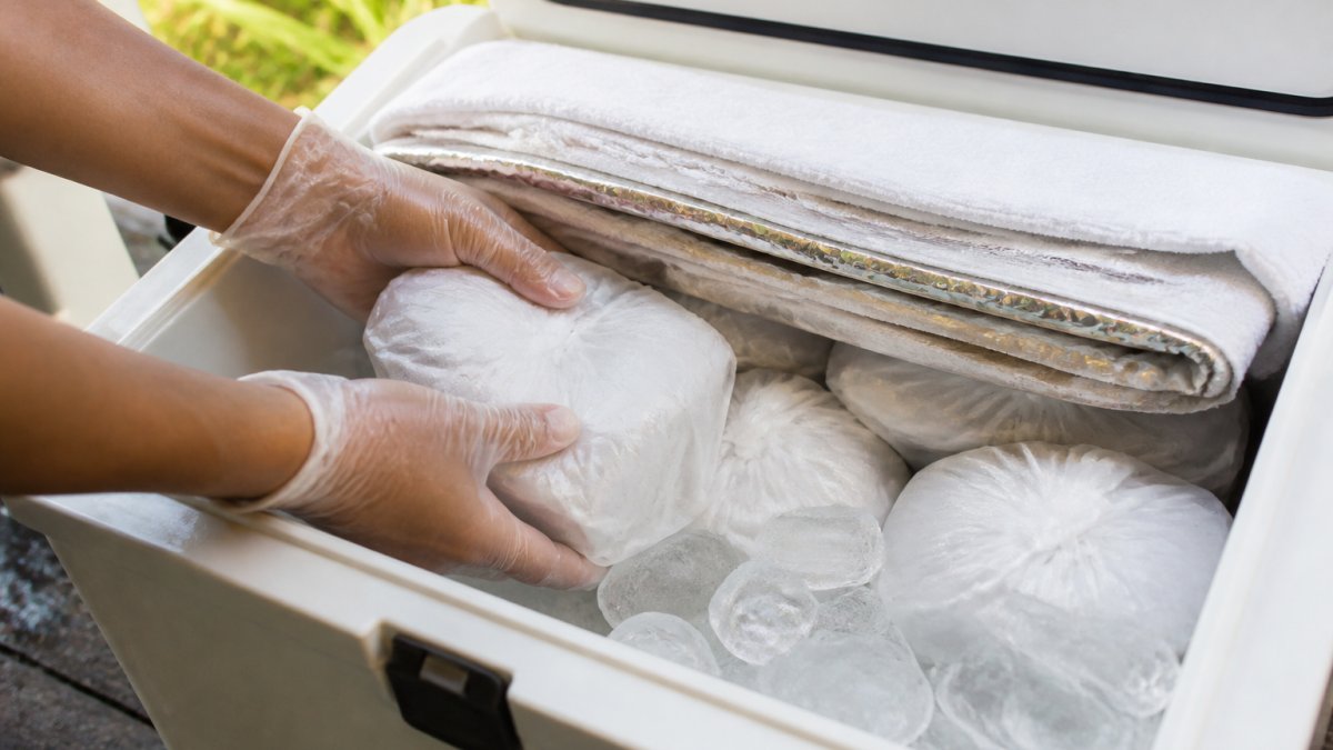 Close-up of gloved hands layering wrapped frozen dessert containers over block ice inside a hard-sided cooler, with a folded towel and foil visible at the top, bright daylight