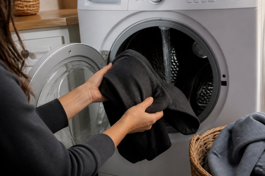 Close-up of hands turning a graphic tee inside-out before placing it into a washing machine, with cold water visible running into the drum
