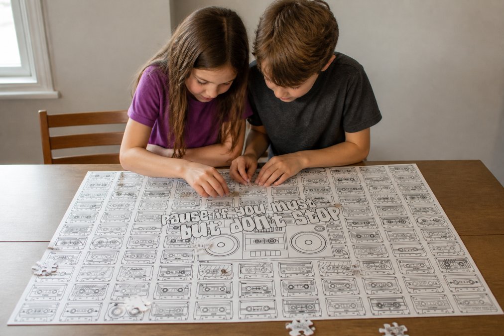 Two siblings working together on a large puzzle at a wooden table, leaning toward each other with focused expressions and collaborative body language, with puzzle pieces scattered across the surface