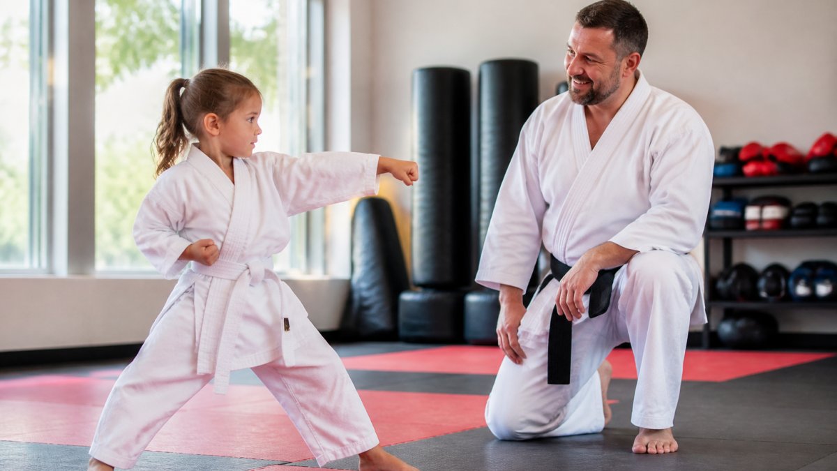 Young child in martial arts uniform practicing a technique with an instructor observing and smiling encouragingly in a bright, welcoming studio with natural light