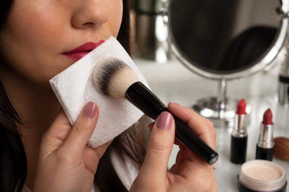 Close-up of a fluffy makeup brush dusting translucent setting powder over a tissue held against lips, with a mirror and lipstick tubes softly blurred in the background