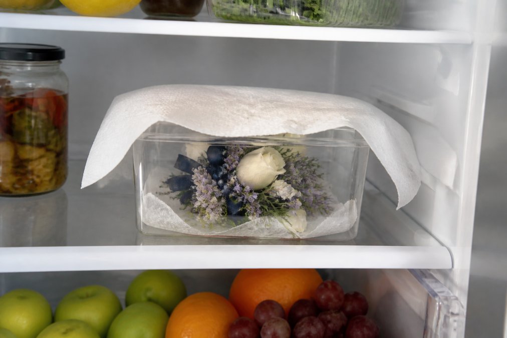 A corsage in a clear plastic box resting on a middle refrigerator shelf, a folded damp paper towel draped over the blooms, fresh fruit visible on a lower shelf below