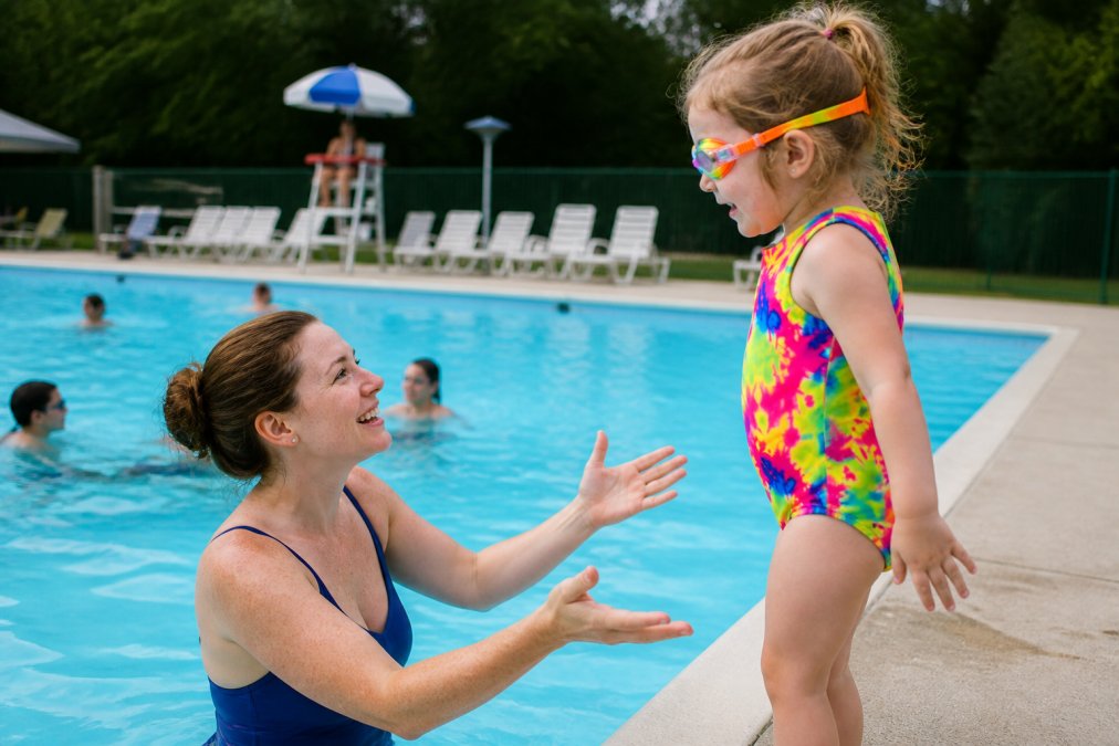 Parent maintaining close visual contact with young toddler at pool's edge during public pool session, demonstrating arm's-reach supervision positioning