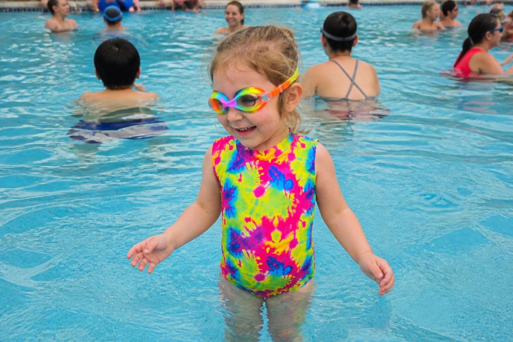 Toddler wearing bright neon yellow swimsuit standing in public pool surrounded by other swimmers, demonstrating how high-visibility color stands out against blue water and other children