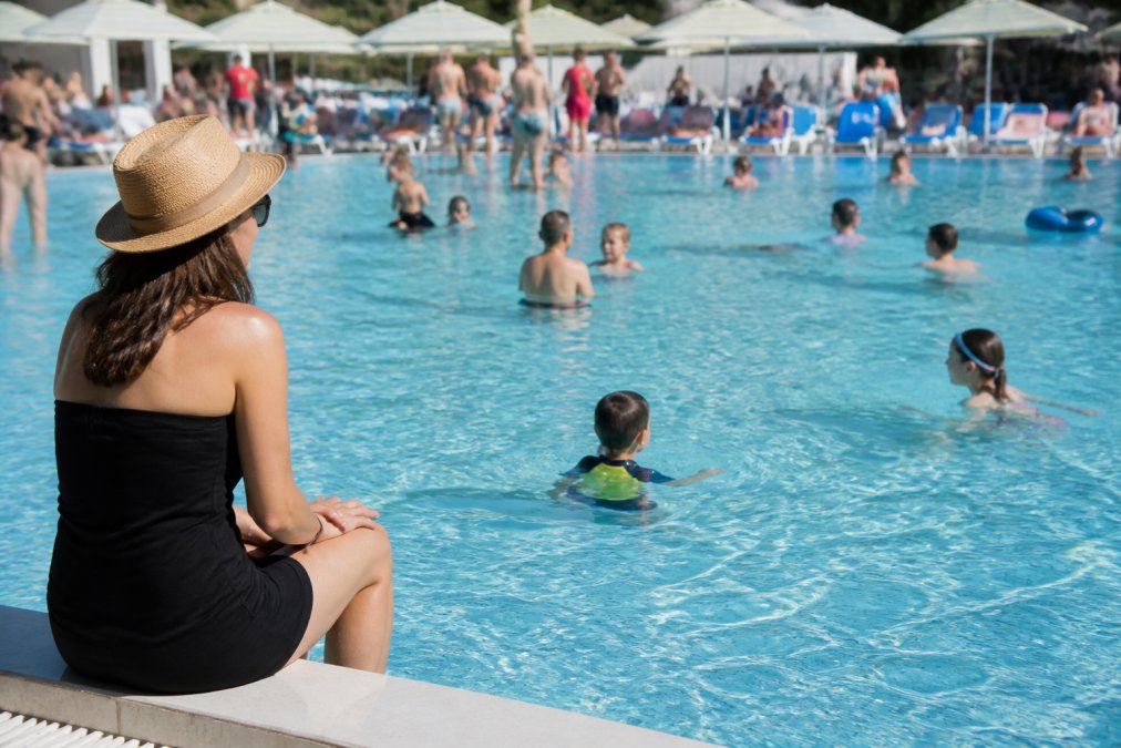 Parent seated poolside at the edge of the pool with clear, direct sightline to the water, watching children swimming while maintaining constant visual focus