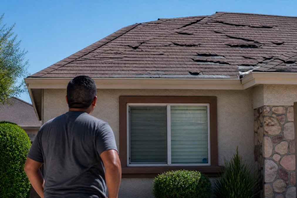 A homeowner standing in their front yard looking up at an aging residential roof with visible missing and curling shingles, clear blue sky, suburban El Paso neighborhood