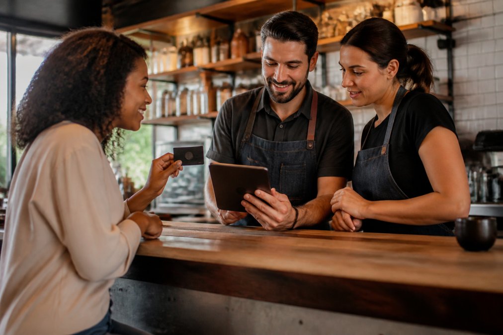 Restaurant owner and manager reviewing membership program details on a tablet at the counter, discussing tiers and benefits with a customer holding a membership card