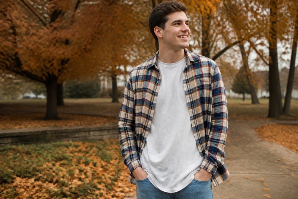 A young adult wearing a faith-based graphic tee under an open flannel shirt, standing outdoors on a crisp fall day with leaves in the background