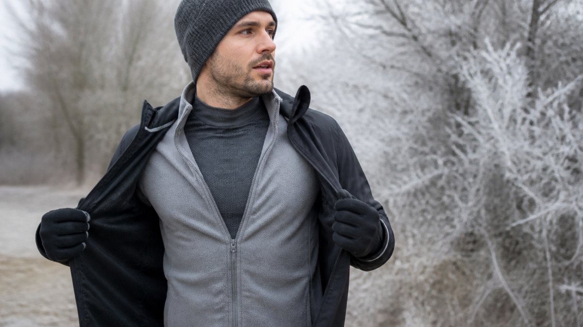 Runner in winter gear standing outdoors showing all three layers clearly visible: fitted base layer under mid-layer fleece under outer windproof jacket, with frost-covered trees in background