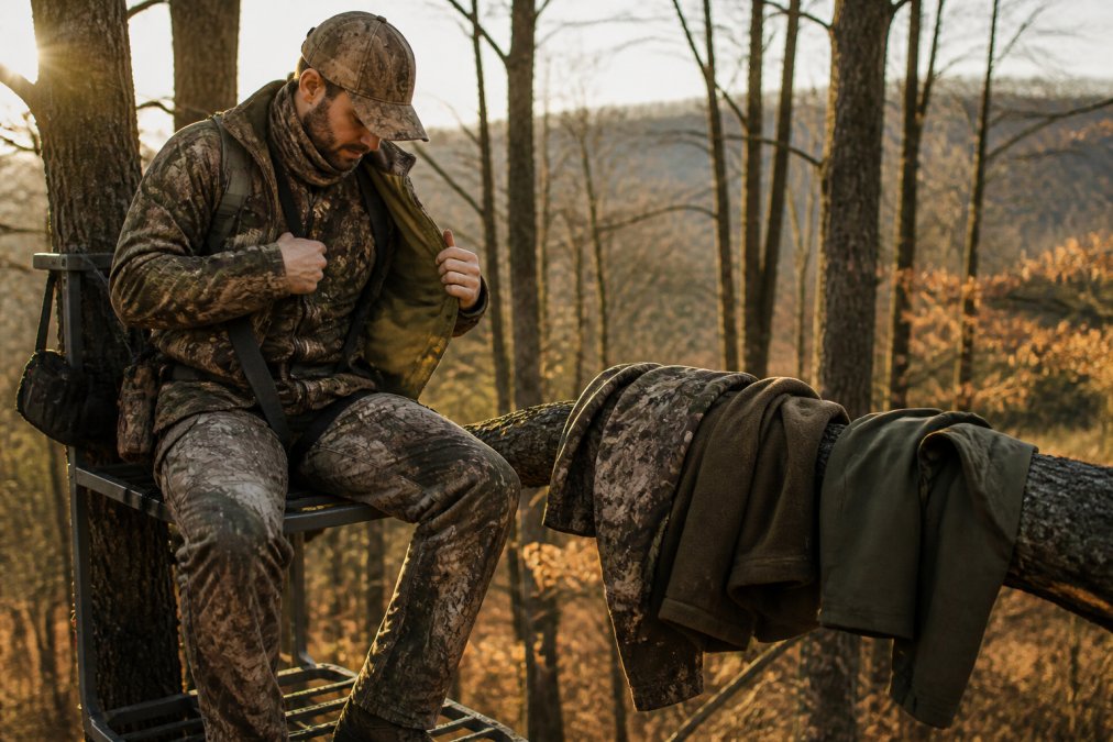A hunter in a tree stand checking layered clothing, with a base layer, mid layer fleece, and outer shell jacket laid out on a nearby branch, early morning light in a wooded area