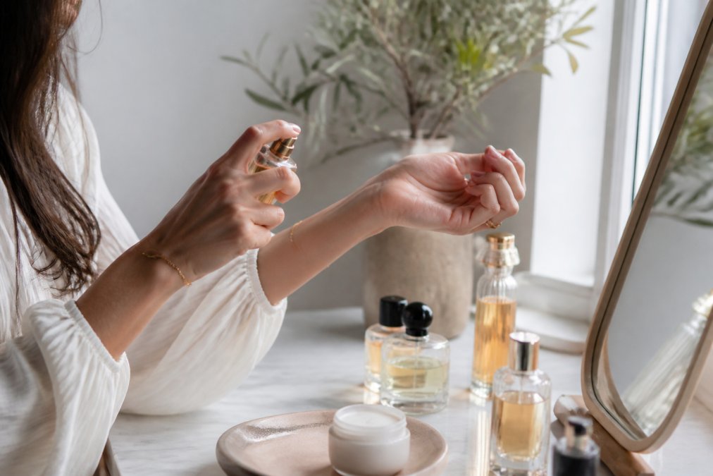 A woman's hands applying perfume to her wrist at a vanity table with several glass fragrance bottles, a small dish of lotion, and soft natural light from a nearby window