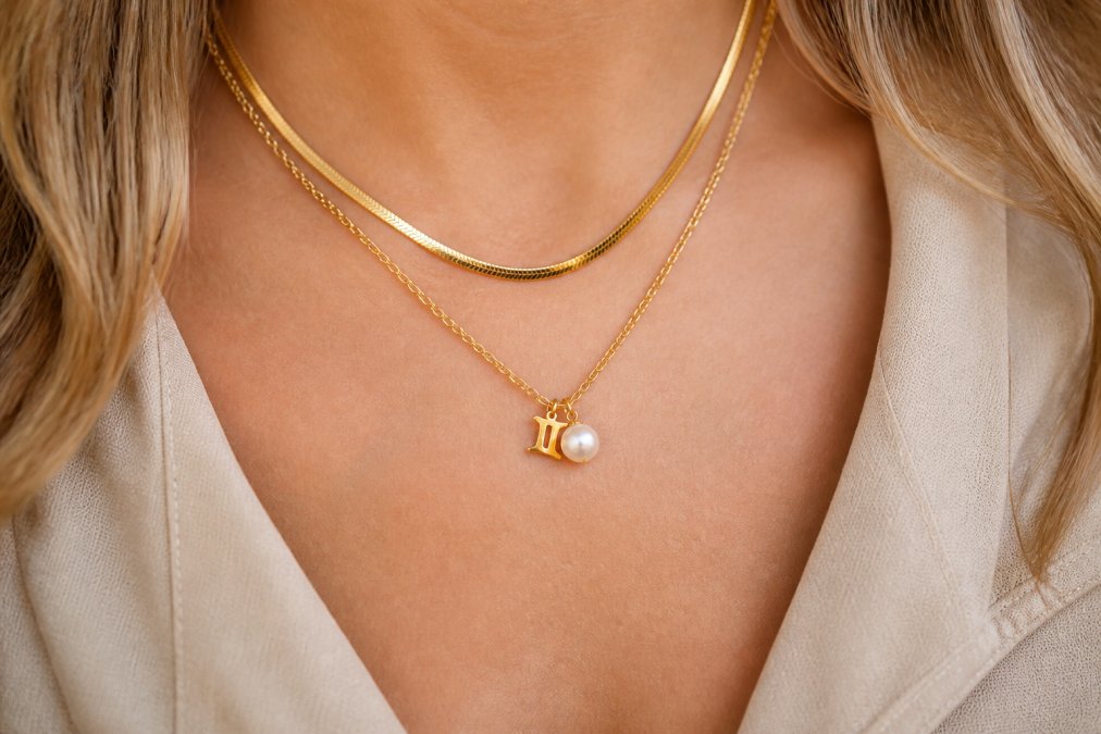 Close-up of a woman's neckline showing a delicate freshwater pearl pendant on a thin gold chain layered beneath a slightly longer, chunkier gold herringbone chain, demonstrating how soft pearl lustre contrasts beautifully with the warm shimmer of gold