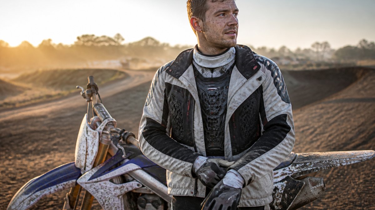 Motocross rider fully geared up standing next to dirt bike on track, showing base layer visible at wrists and neck, protective textile jacket, and armor pieces, with dusty track and morning light in background