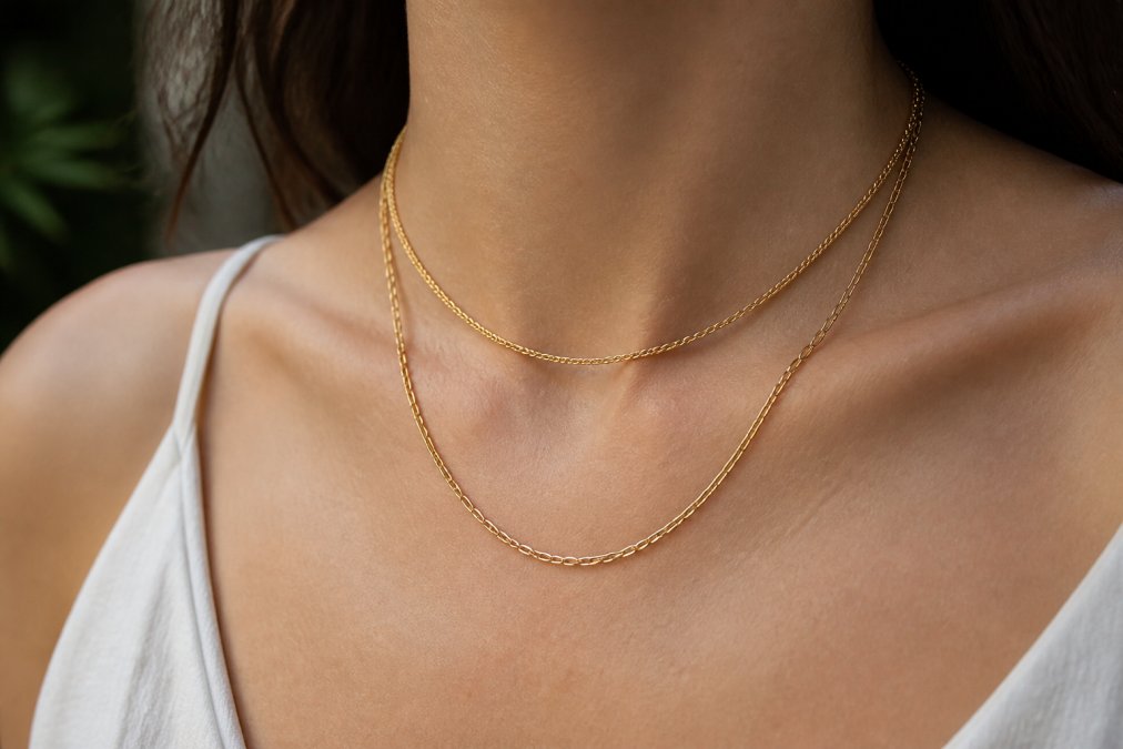 Close-up of a woman's neck and collarbone wearing two layered gold chains of different lengths, with the shorter chain sitting higher and the longer chain draping lower, natural daylight highlighting the texture and finish of the metals