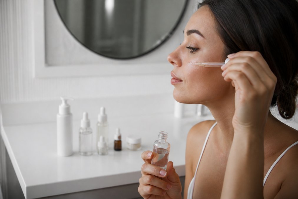 Woman applying serum to her cheek with chemical peel skincare bottles lined up on a clean bathroom counter
