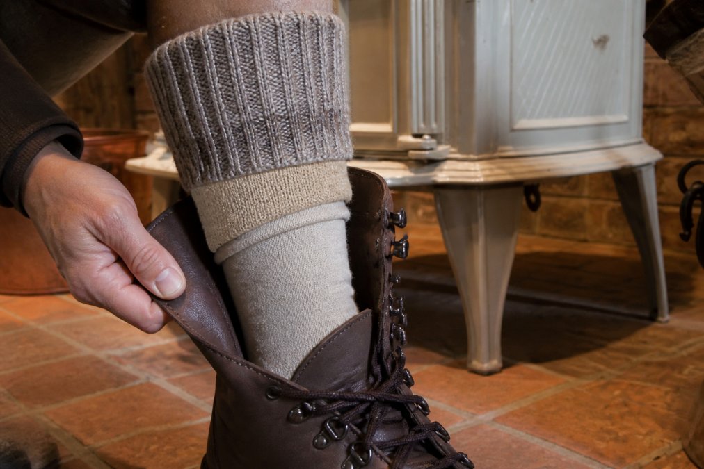Close-up of feet wearing properly layered socks inside a winter boot, showing the thin base layer against skin, mid-weight insulating sock in the middle, and heavier alpaca outer sock visible at the calf
