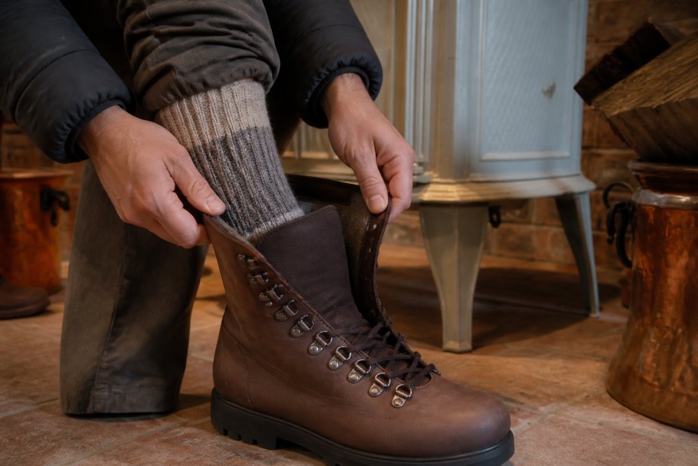 Hands adjusting a winter hiking boot with proper toe box space, demonstrating correct fit with layered alpaca socks visible at the calf, showing the gap between toes and boot interior