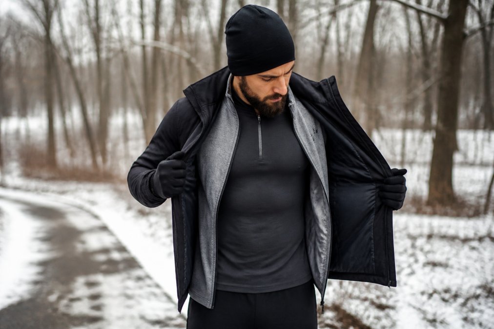 A person outdoors in cold weather wearing a visible three-layer winter workout outfit, peeling back the outer shell to reveal the mid-layer and base layer underneath, with snow-dusted ground and bare trees in the background