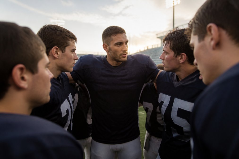 Team captain standing shoulder-to-shoulder with teammates during pre-game huddle, maintaining composed posture while making direct eye contact with each player, natural stadium lighting in background