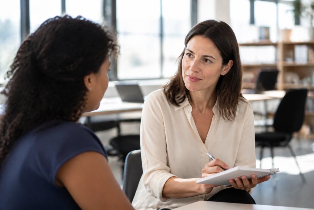 A female manager in a bright office leaning forward and making eye contact with a colleague during a one-on-one conversation, holding a notebook