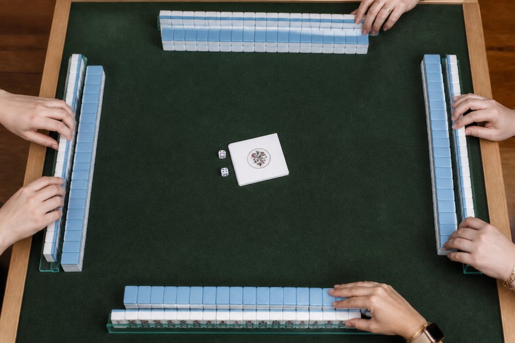 Overhead shot of a mahjong table with four racks of tiles arranged in neat rows, dice resting in the center beside a folded NMJL card, players' hands adjusting tiles under warm indoor lighting
