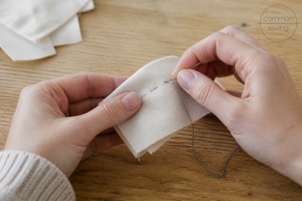 Close-up of hands holding a needle and thread over a folded fabric scrap, showing a running stitch in progress on a plain wooden worktable in soft daylight