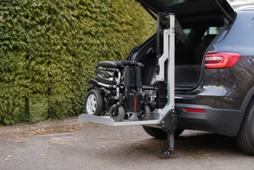 Close-up view of a portable electric car boot hoist mechanism in operation, smoothly raising a folded powerchair toward an open vehicle boot with the lifting platform clearly visible