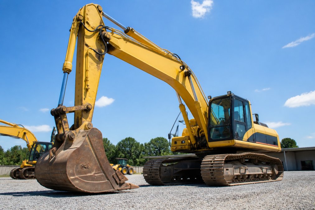 Heavy excavator equipment photographed in bright daylight, showing clear details of the bucket, boom, and hydraulic systems from multiple angles, with shadows creating depth and definition