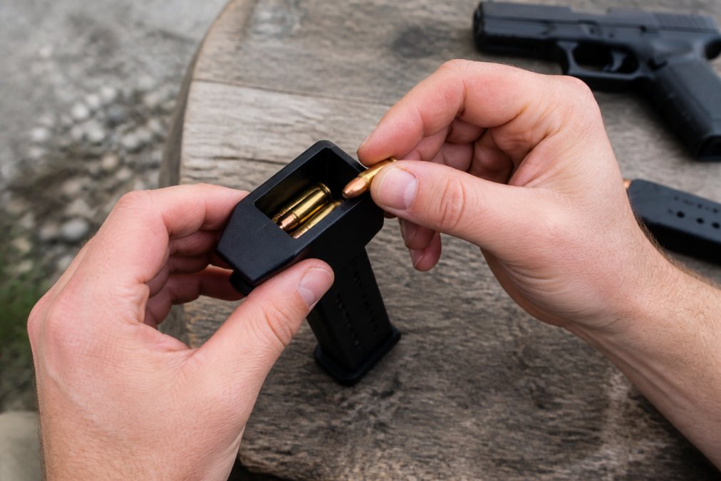 Hands inserting 9mm magazine into speed loader device with one hand while feeding cartridges into the open magazine top with the other hand, showing the loading mechanism in action