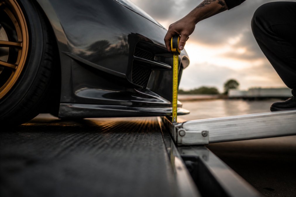 Close-up of a lowered sports car's front splitter and undercarriage positioned near a trailer ramp, showing the tight clearance between the body and the loading surface with precise measurement markers visible