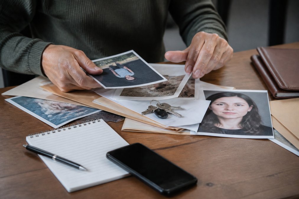 Close-up of hands organizing missing person documents, photographs, and personal items on a wooden table with notepad, pen, and smartphone