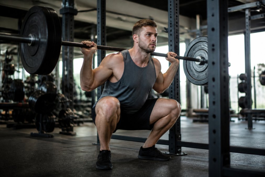 Athletic individual performing a barbell squat in a well-lit gym with proper form and intense focus, demonstrating controlled descent