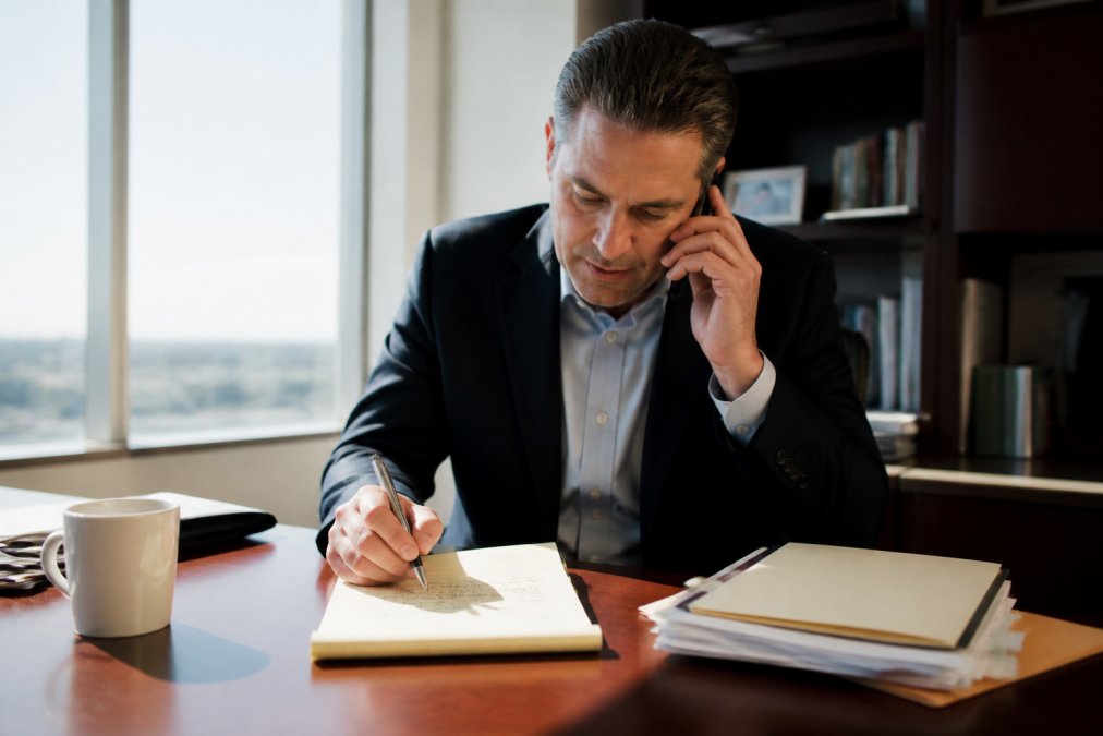 A business professional sitting at an office desk during a phone call, notepad open with handwritten bullet points in front of them, confident posture, natural window light