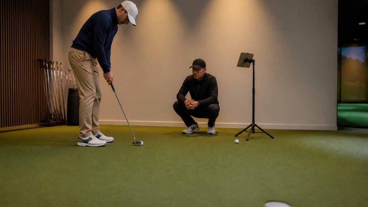 A golfer practicing a putting stroke on an indoor green while a coach watches and a tablet on a stand shows shot data nearby, warm overhead lighting