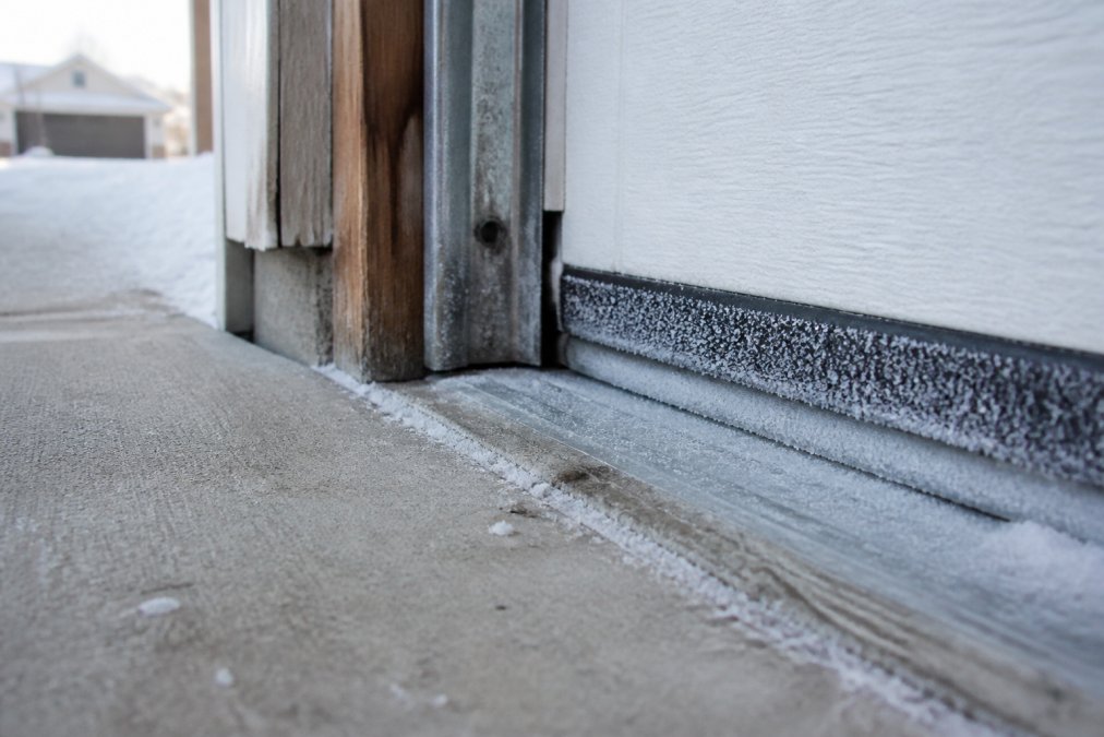 Garage door bottom seal and concrete threshold in winter conditions with frost visible on weatherstripping and proper drainage slope away from the door
