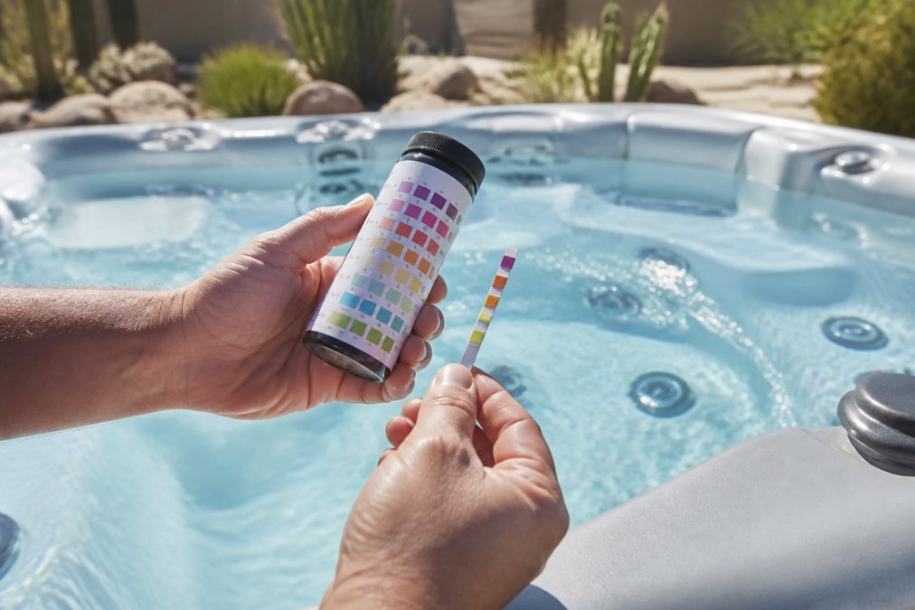 A close-up of a hand holding a water test strip over an open spa, comparing the colored pads to a reference chart on a plastic bottle, with bright desert sunlight sparkling on the water surface