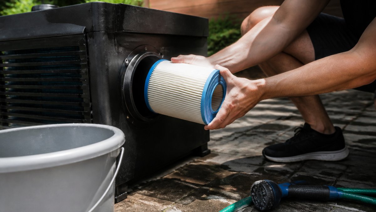 Close-up of hands removing a pleated filter cartridge from a cold plunge chiller housing, with a garden hose and bucket nearby on a patio