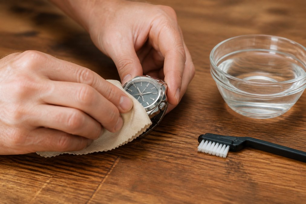 A mechanical watch being gently wiped with a lint-free cloth on a clean wooden surface, with a soft brush and a small bowl of water visible beside it