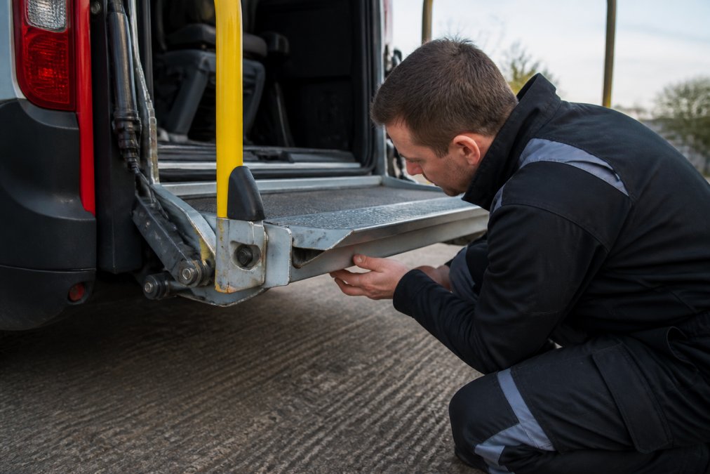 Professional maintenance technician performing close-up visual inspection of minibus tail lift platform, checking for corrosion, loose bolts, and hydraulic fluid connections, with vehicle positioned on flat ground and morning natural lighting