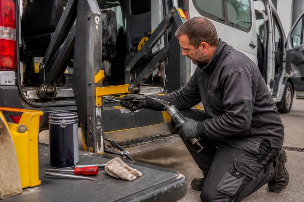 Experienced maintenance technician applying grease to the pivot hinges and mechanical joints of a minibus tail lift mechanism, with grease gun in hand and lubrication supplies arranged on a work surface, vehicle on flat ground