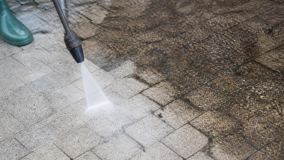 Close-up of block paving driveway being cleaned with a pressure washer, showing the contrast between freshly cleaned light grey blocks and darker stained sections, with water spray visible on the surface