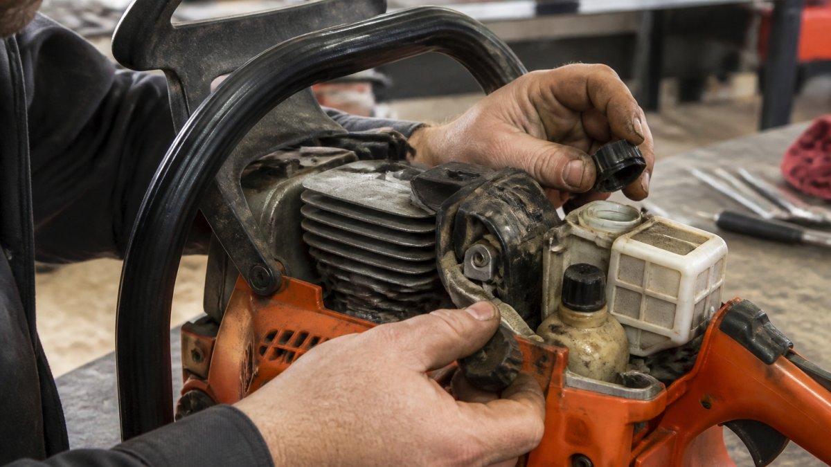 Professional mechanic's hands inspecting chainsaw engine components, checking fuel cap and oil cap with close-up detail of cooling fins on aluminum engine block in well-lit workshop