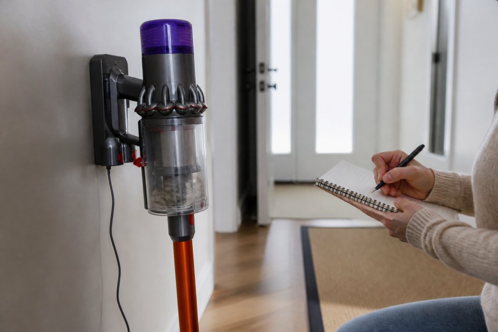 Close-up of a cordless stick vacuum resting on its charging dock in a bright hallway, with a hand holding a small notebook and pen in the background, suggesting a homeowner tracking battery performance over time