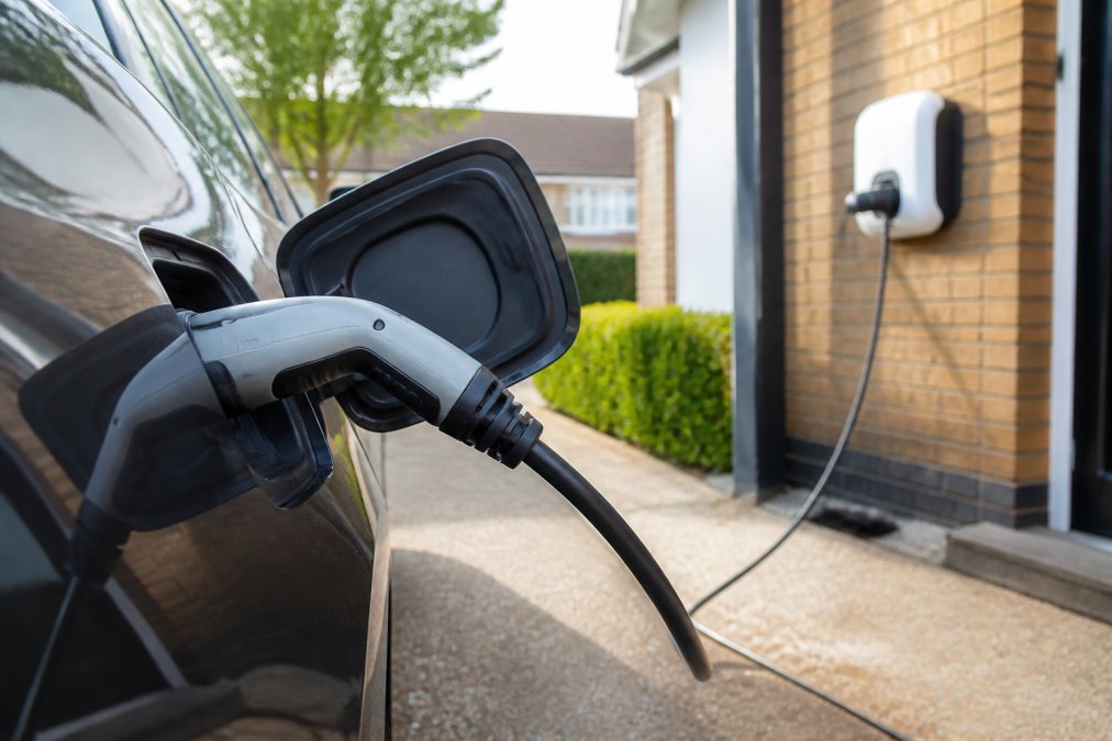 Close-up of an electric vehicle charging cable connected to a car's charging port, with a home charging station visible in the background during daytime