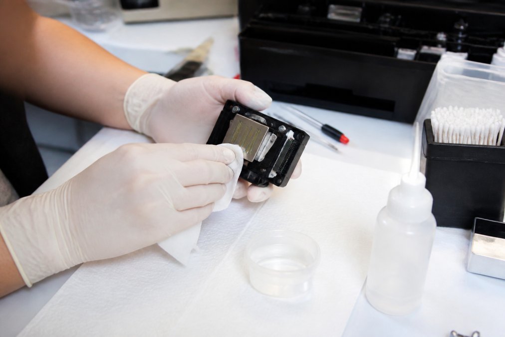 Close-up of a person's hands wearing latex gloves, carefully using a lint-free cloth and a small container of cleaning solution to wipe the bottom of an Epson printhead removed from a printer on a clean, well-lit workbench