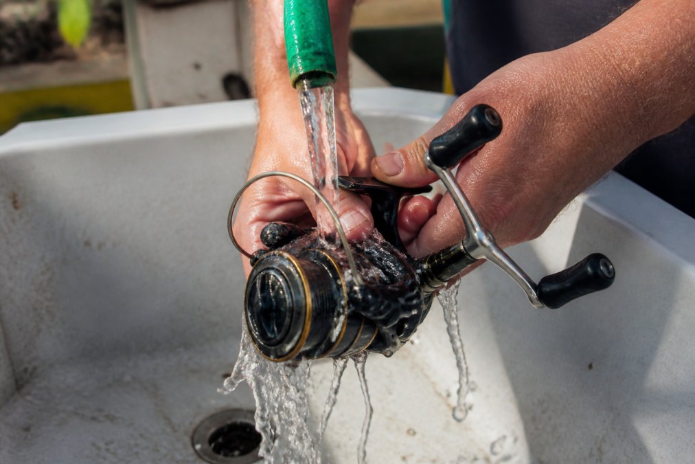 Close-up of hands rinsing a fishing reel under freshwater from a hose, showing water flowing over the reel mechanism and handles with salt residue washing away